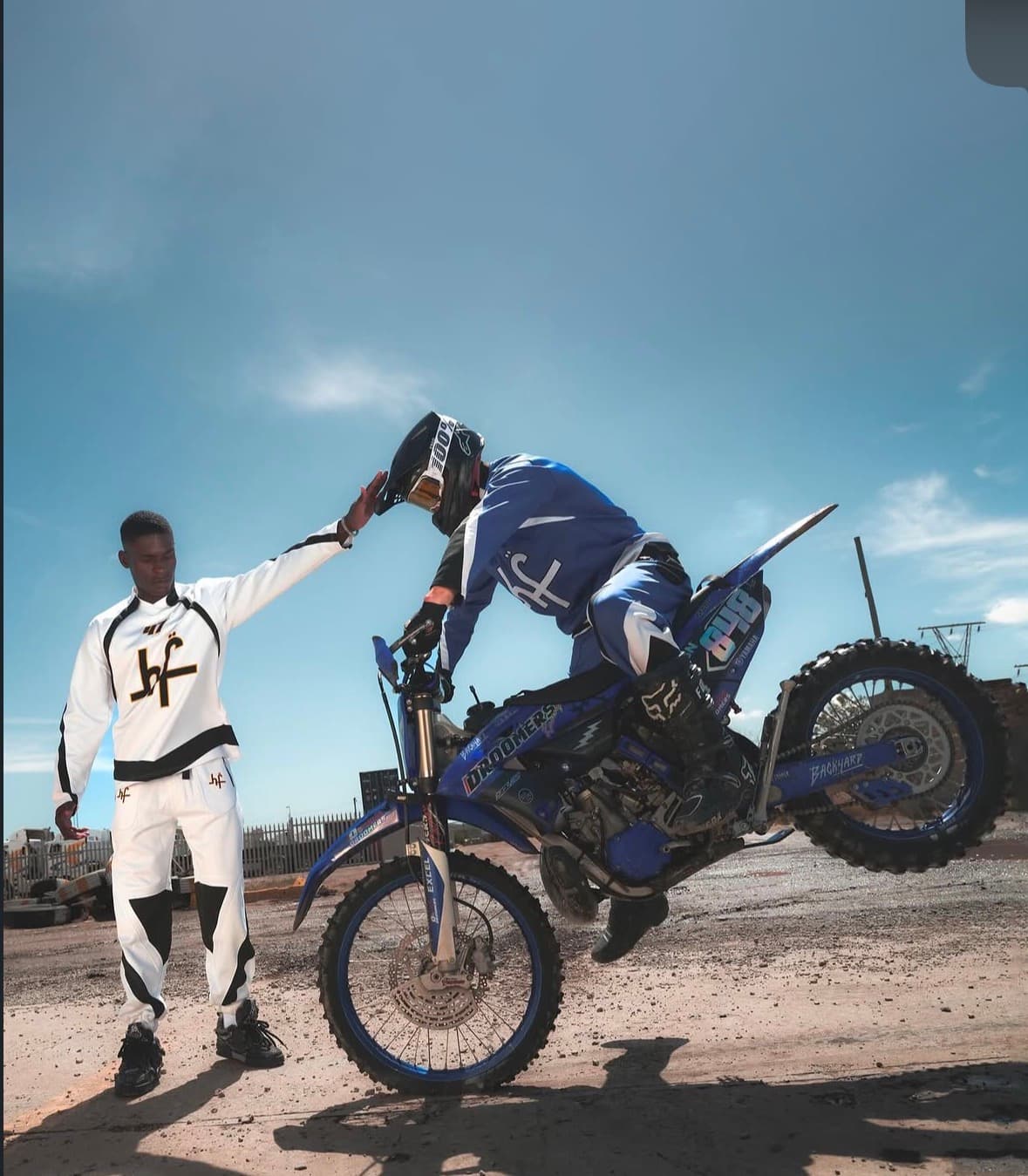 Man in white gear touches the helmet of a motocross rider performing a nose wheelie.