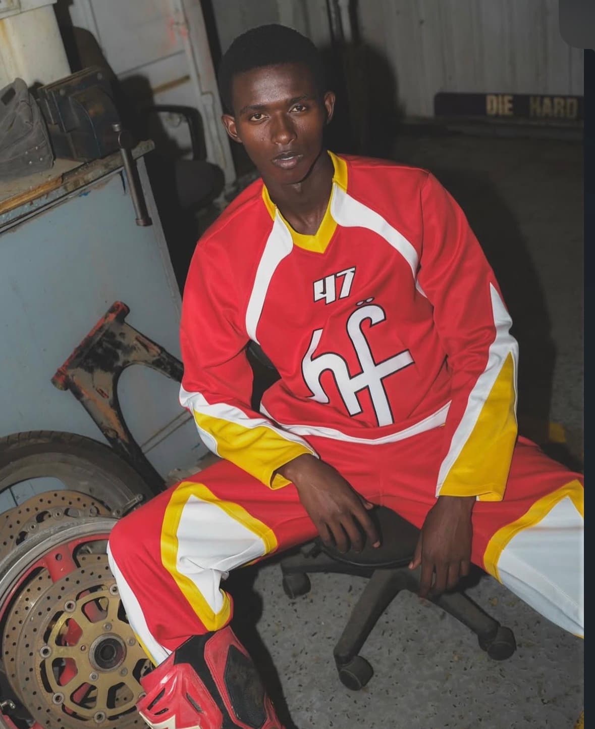 Young man in a red and yellow racing suit sits in a cluttered motorcycle workshop.