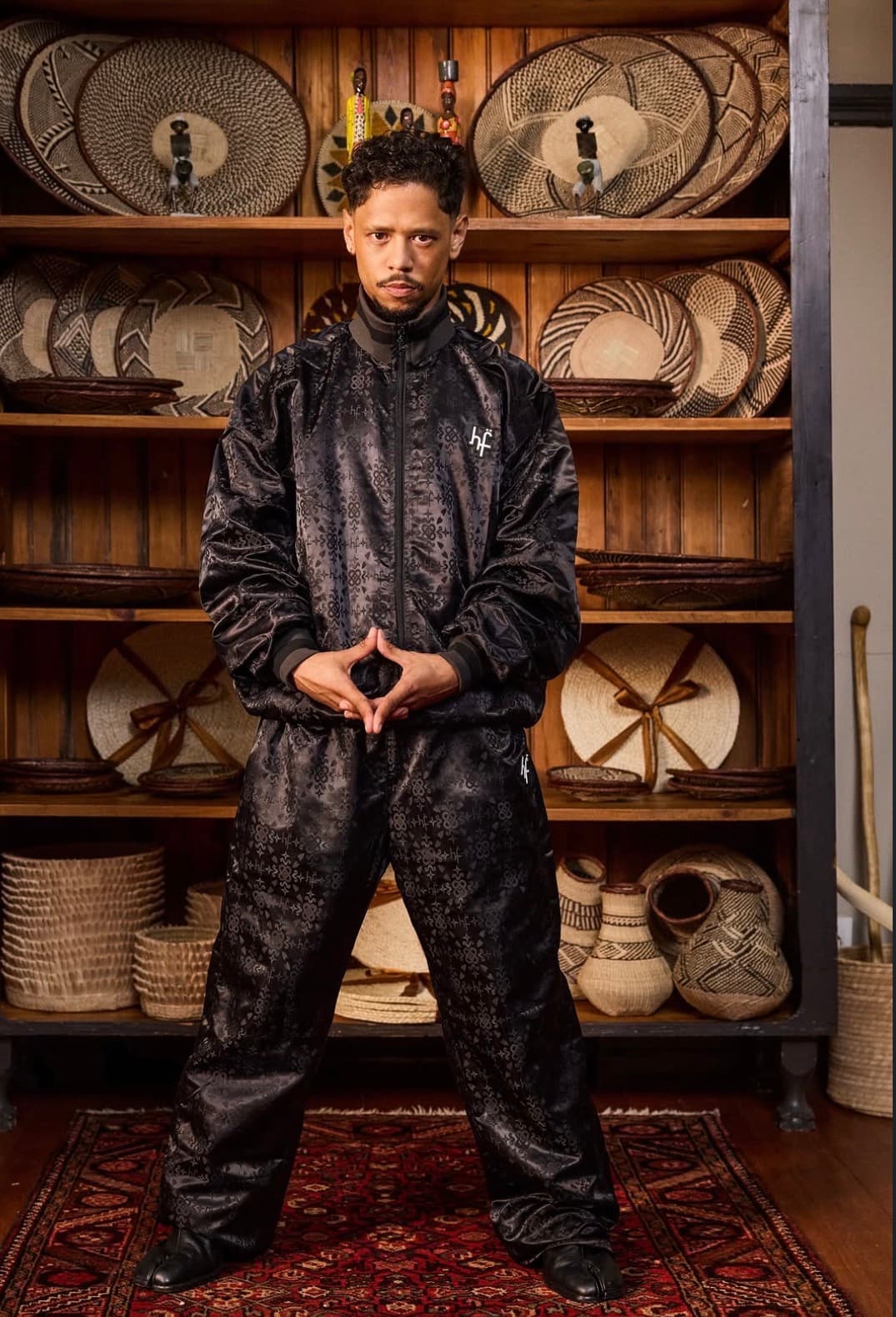 Man in black patterned tracksuit posing before shelves of traditional woven baskets and plates.