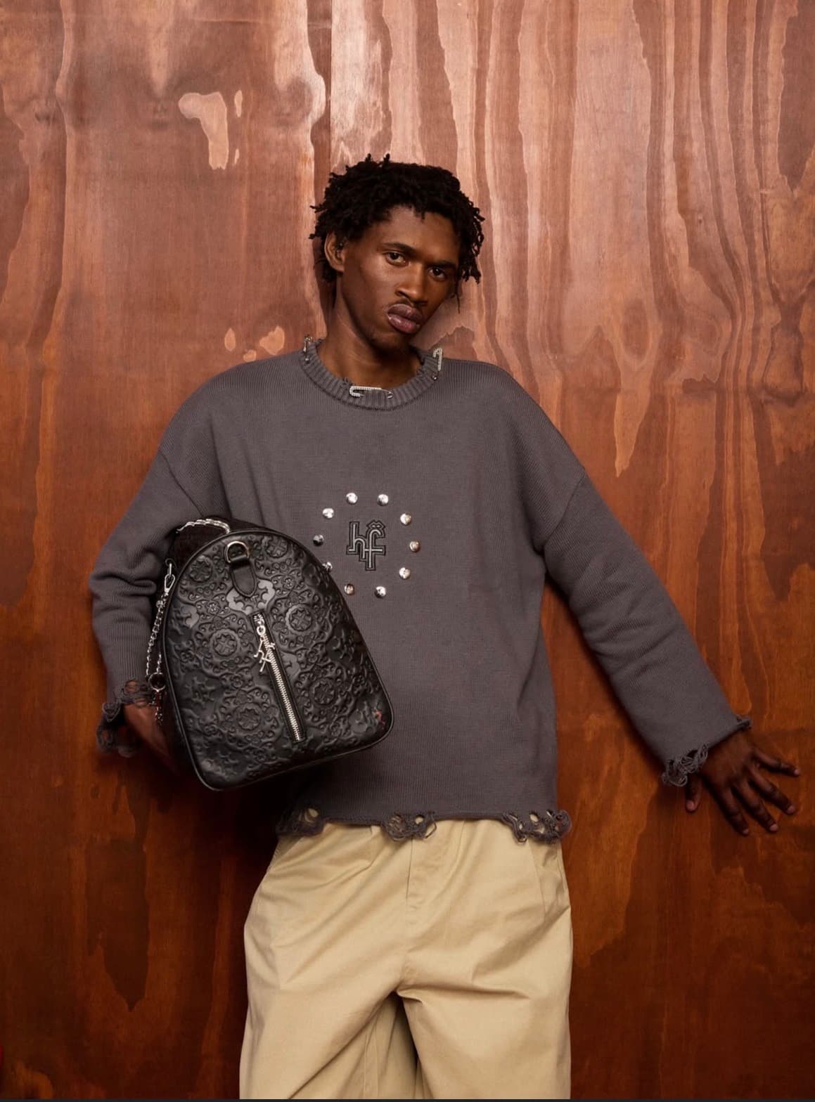 Man in a grey distressed sweater holding a black embossed leather bag against wood paneling.