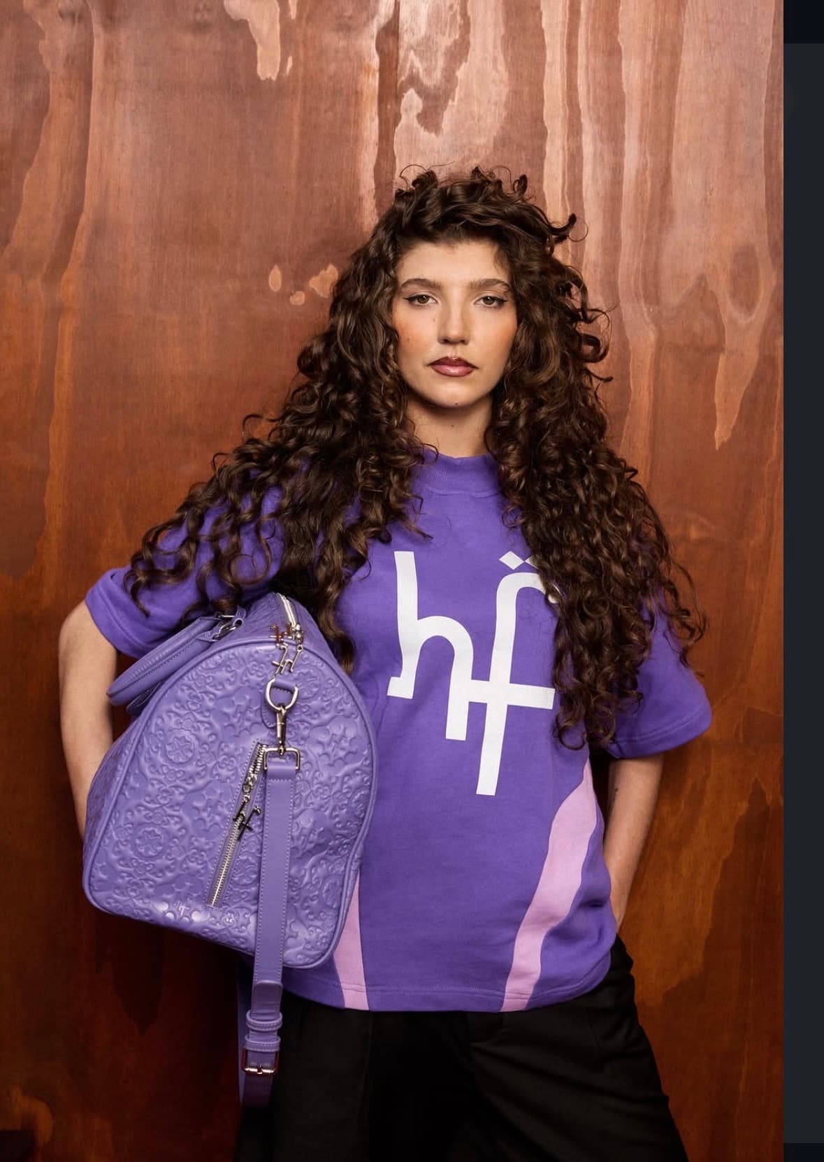 Curly-haired woman in a purple logo t-shirt holding a matching embossed purple duffle bag.